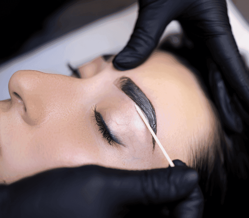 Person receiving eyebrow tinting treatment with a wooden stick, hands in black gloves.