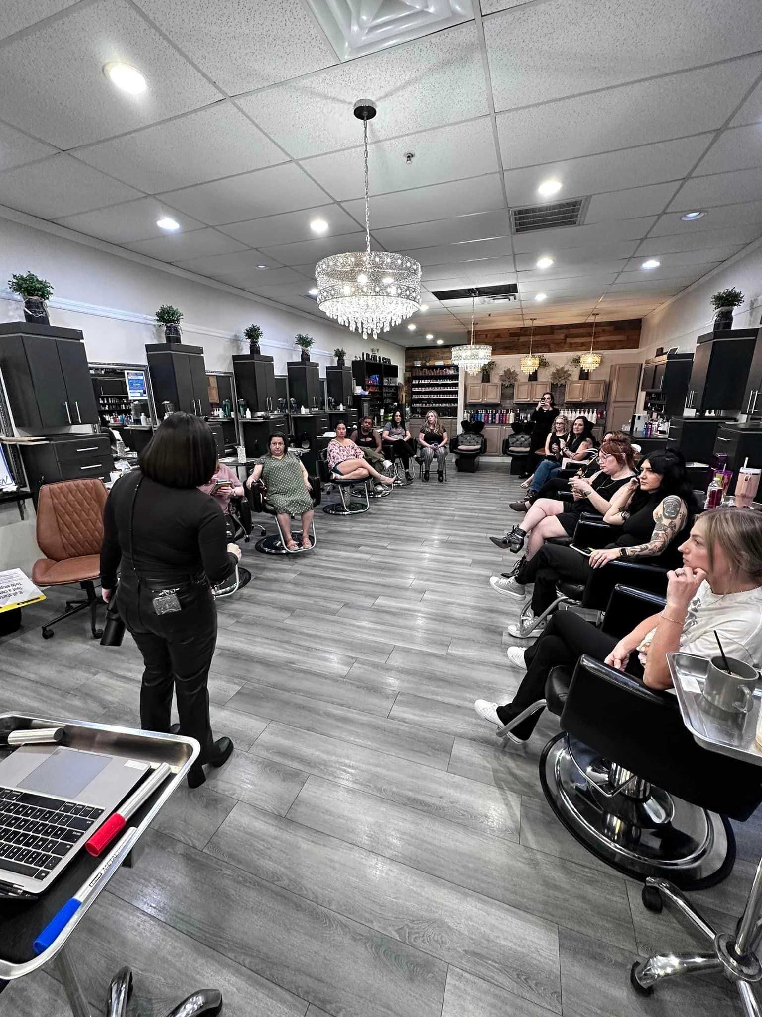 A woman presents to a group seated in a modern salon with chandeliers and black chairs.