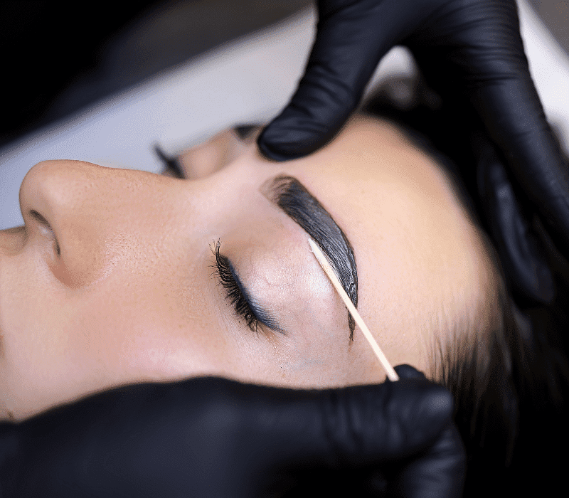 Person receiving eyebrow tinting treatment with a wooden stick, hands in black gloves.